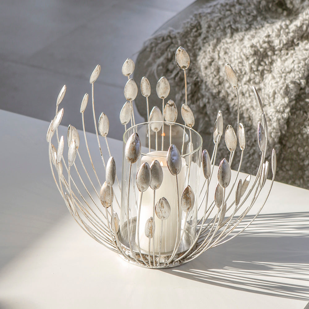 Das Windlicht "Trevi" ist aus Metall gefertigt und in einem antik-silbernen Design gefärbt. Auf der runden Basis befindet sich ein Glaseinsatz, umgeben von natürlich anmutenden Blüten- oder Blumenstauden.