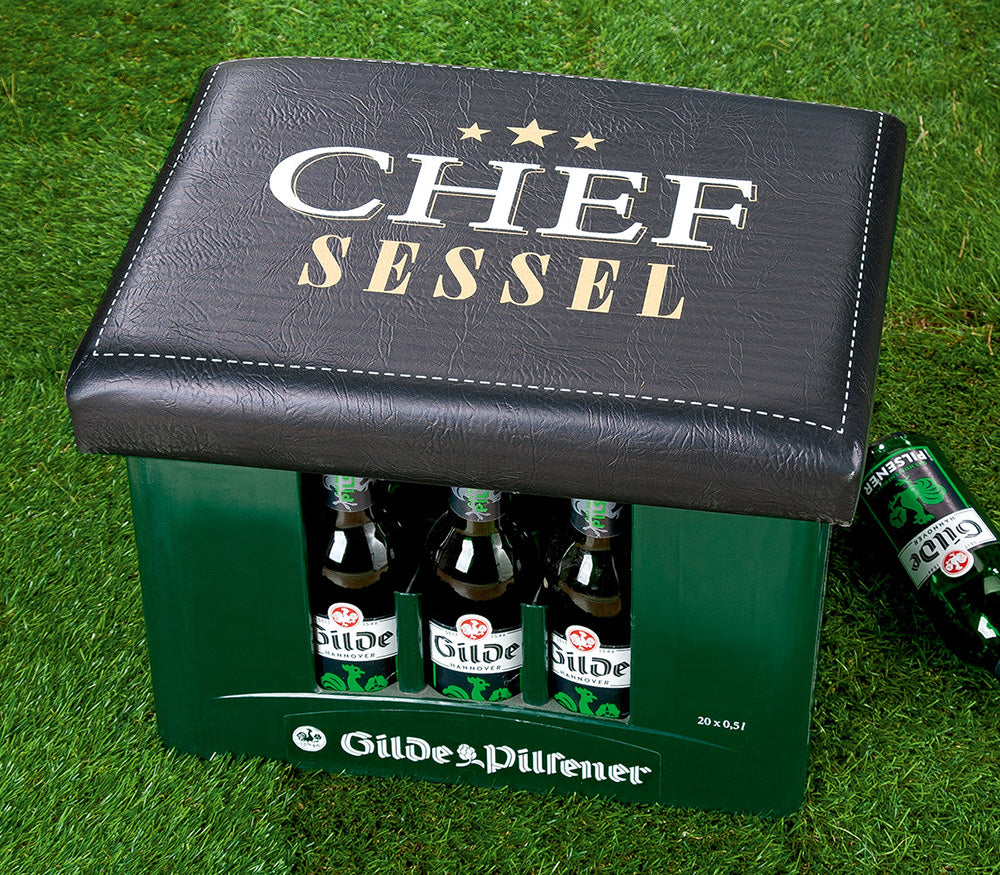 Sitzpolster Chef Sessel für Bierkisten. Schwarz mit Aufschrift Chef Sessel auf der Oberseite. Dieses praktische und originelle Kissen lässt sich einfach auf jede Bierkiste legen und verwandelt sie in einen bequemen Sitzplatz. Abbildung mit Bierkasten

