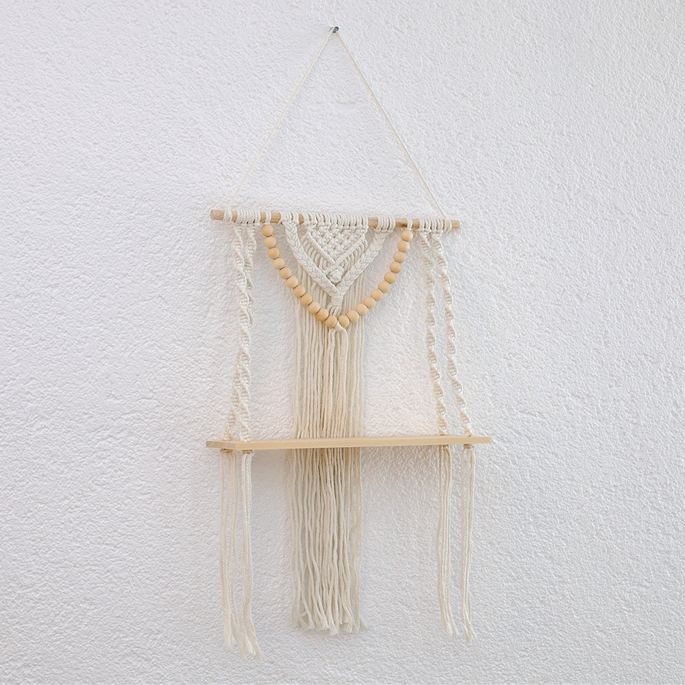 DECCARO Makramee-Hängeregal Bead. Vertikale Holzstange an der Baumwollseile befestigt sind, die mit einem stabilen, schmalen Holzbrett verknüpft sind. Weiterhin ist an der Holzstange ein Makramee Knotenmuster mit vielen kleinen Holzperlen befestigt, die die Optik des Regals abrunden.