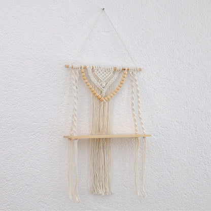 DECCARO Makramee-Hängeregal Bead. Vertikale Holzstange an der Baumwollseile befestigt sind, die mit einem stabilen, schmalen Holzbrett verknüpft sind. Weiterhin ist an der Holzstange ein Makramee Knotenmuster mit vielen kleinen Holzperlen befestigt, die die Optik des Regals abrunden.